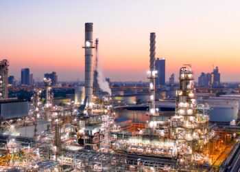 An extensive industrial refinery complex is shown at dusk, with several tall chimneys and metal structures. The site, reminiscent of a thriving gush of stock activity, is illuminated with bright lights. In the background, an urban skyline is visible with high-rise buildings against a colorful sunset sky. | 5StarsStocks