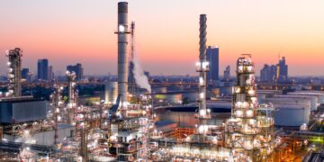 An extensive industrial refinery complex is shown at dusk, with several tall chimneys and metal structures. The site, reminiscent of a thriving gush of stock activity, is illuminated with bright lights. In the background, an urban skyline is visible with high-rise buildings against a colorful sunset sky. | 5StarsStocks