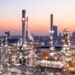 An extensive industrial refinery complex is shown at dusk, with several tall chimneys and metal structures. The site, reminiscent of a thriving gush of stock activity, is illuminated with bright lights. In the background, an urban skyline is visible with high-rise buildings against a colorful sunset sky. | 5StarsStocks