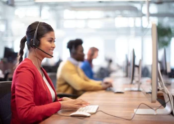 A woman in a red jacket sits at a desk, wearing a headset and typing on a keyboard in a modern, open office. Behind her, other colleagues also wearing headsets work diligently at their computers, perhaps discussing the latest fluctuations in SPRT Stock. The room is bright with natural light. | 5StarsStocks