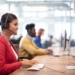 A woman in a red jacket sits at a desk, wearing a headset and typing on a keyboard in a modern, open office. Behind her, other colleagues also wearing headsets work diligently at their computers, perhaps discussing the latest fluctuations in SPRT Stock. The room is bright with natural light. | 5StarsStocks