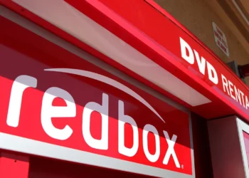 Close-up view of a redbox DVD rental kiosk. The bold red cabinet has the word "redbox" prominently displayed in white text, along with "DVD RENTALS" above it. The kiosk is situated outdoors against a beige wall, with some daylight visible—an everyday sight for investors keeping an eye on RDBX stock. | 5StarsStocks