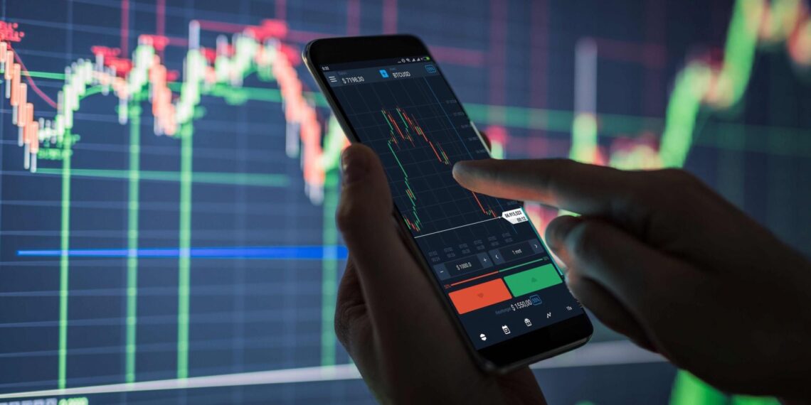 A person is holding a smartphone and analyzing stock market data displayed on the screen. A large monitor in the background shows colorful financial graphs and charts from 5StarsStocks, indicating stock market trends. The scene suggests active trading or financial analysis of Stocks to Invest. | 5StarsStocks