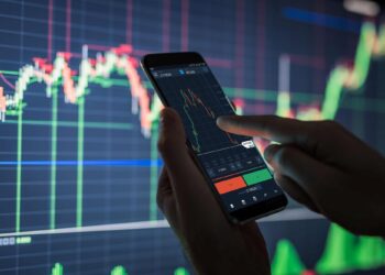 A person is holding a smartphone and analyzing stock market data displayed on the screen. A large monitor in the background shows colorful financial graphs and charts from 5StarsStocks, indicating stock market trends. The scene suggests active trading or financial analysis of Stocks to Invest. | 5StarsStocks