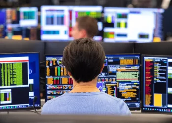 A person sits in front of multiple computer monitors displaying various financial data and charts, analyzing 5starsstocks. The background shows more monitors and other people working in what appears to be a busy trading room or financial office, all focused on identifying the best stocks to invest. | 5StarsStocks