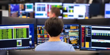 A person sits in front of multiple computer monitors displaying various financial data and charts, analyzing 5starsstocks. The background shows more monitors and other people working in what appears to be a busy trading room or financial office, all focused on identifying the best stocks to invest. | 5StarsStocks