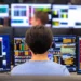 A person sits in front of multiple computer monitors displaying various financial data and charts, analyzing 5starsstocks. The background shows more monitors and other people working in what appears to be a busy trading room or financial office, all focused on identifying the best stocks to invest. | 5StarsStocks