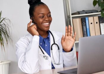 A doctor in a white coat with a stethoscope around their neck is sitting at a desk, smiling and waving at a laptop. They are wearing a headset, suggesting a telehealth consultation. A potted plant and a bookshelf with binders and medical stocks are in the background. | 5StarsStocks