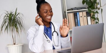 A doctor in a white coat with a stethoscope around their neck is sitting at a desk, smiling and waving at a laptop. They are wearing a headset, suggesting a telehealth consultation. A potted plant and a bookshelf with binders and medical stocks are in the background. | 5StarsStocks