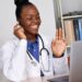 A doctor in a white coat with a stethoscope around their neck is sitting at a desk, smiling and waving at a laptop. They are wearing a headset, suggesting a telehealth consultation. A potted plant and a bookshelf with binders and medical stocks are in the background. | 5StarsStocks