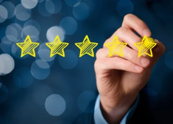 A hand holding a yellow marker is drawing five yellow stars in a row against a dark, blurred background with blue bokeh lights. The image represents 5StarsStocks and signifies the concept of high quality or top-tier performance. | 5StarsStocks