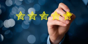 A hand holding a yellow marker is drawing five yellow stars in a row against a dark, blurred background with blue bokeh lights. The image represents 5StarsStocks and signifies the concept of high quality or top-tier performance. | 5StarsStocks