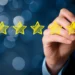 A hand holding a yellow marker is drawing five yellow stars in a row against a dark, blurred background with blue bokeh lights. The image represents 5StarsStocks and signifies the concept of high quality or top-tier performance. | 5StarsStocks