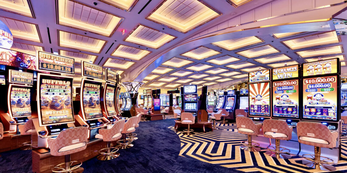 A brightly lit casino floor with rows of colorful slot machines and video poker games creates a vivacious scene akin to thriving casino stocks. The ceiling features a modern, geometric design with recessed lighting. Several white and gold cushioned chairs are positioned at the machines, crafting an inviting atmosphere for players. | 5StarsStocks