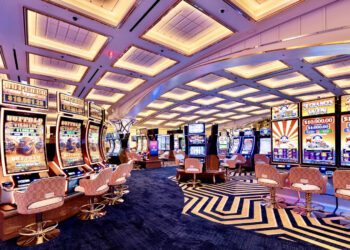 A brightly lit casino floor with rows of colorful slot machines and video poker games creates a vivacious scene akin to thriving casino stocks. The ceiling features a modern, geometric design with recessed lighting. Several white and gold cushioned chairs are positioned at the machines, crafting an inviting atmosphere for players. | 5StarsStocks
