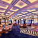 A brightly lit casino floor with rows of colorful slot machines and video poker games creates a vivacious scene akin to thriving casino stocks. The ceiling features a modern, geometric design with recessed lighting. Several white and gold cushioned chairs are positioned at the machines, crafting an inviting atmosphere for players. | 5StarsStocks