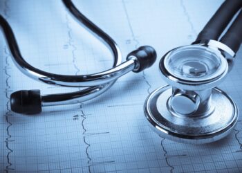 A stethoscope lies on top of an electrocardiogram (ECG or EKG) printout. The stethoscope, with its metal chest piece and flexible tubing, is prominently displayed in the foreground, while the ECG graph, showing heart activity and hinting at trends in health stocks, is visible beneath it. | 5StarsStocks