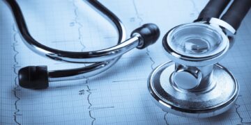 A stethoscope lies on top of an electrocardiogram (ECG or EKG) printout. The stethoscope, with its metal chest piece and flexible tubing, is prominently displayed in the foreground, while the ECG graph, showing heart activity and hinting at trends in health stocks, is visible beneath it. | 5StarsStocks
