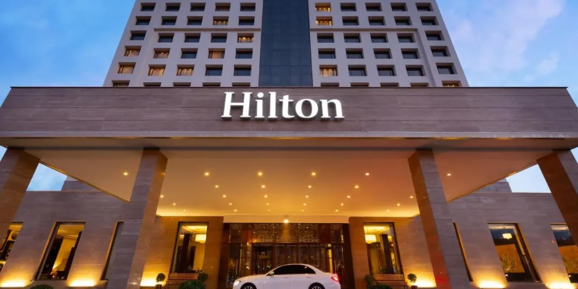 A large hotel building with the "Hilton" logo prominently displayed at the top and above the entrance. The hotel has multiple floors with rows of lit windows, hinting at bustling business activity possibly influencing Hotel Stocks. A white car is parked under a brightly lit, covered driveway at the main entrance. | 5StarsStocks