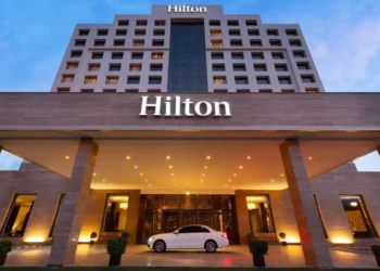 A large hotel building with the "Hilton" logo prominently displayed at the top and above the entrance. The hotel has multiple floors with rows of lit windows, hinting at bustling business activity possibly influencing Hotel Stocks. A white car is parked under a brightly lit, covered driveway at the main entrance. | 5StarsStocks