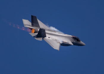 A fighter jet, reminiscent of a top-tier play in military stocks, flies through a clear blue sky, banked to its left side. The aircraft exhibits afterburner flames and condensation trails over its wings, indicating high-speed flight. | 5StarsStocks