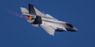 A fighter jet, reminiscent of a top-tier play in military stocks, flies through a clear blue sky, banked to its left side. The aircraft exhibits afterburner flames and condensation trails over its wings, indicating high-speed flight. | 5StarsStocks