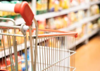 A close-up view of an empty shopping cart with a red handlebar in the aisle of a grocery store. Shelves filled with a variety of staples are slightly blurred in the background. The image focuses on the cart, suggesting a shopping experience and the need to stock up on essentials. | 5StarsStocks