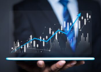 A person in a suit holds a tablet displaying a holographic stock market chart with upward trends. The chart includes candlestick patterns, data points, and an ascending blue arrow indicating growth and positive market performance—ideal for identifying stocks to invest in. | 5StarsStocks