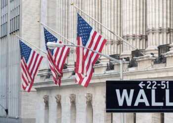 Four American flags fly in front of buildings with columns on Wall Street. A black street sign displaying "Wall St" with arrows pointing to 22-51 is in the foreground. The stone buildings are well-lit with natural light, embodying the financial essence of Wall Street, home of the Dow Jones. | 5StarsStocks
