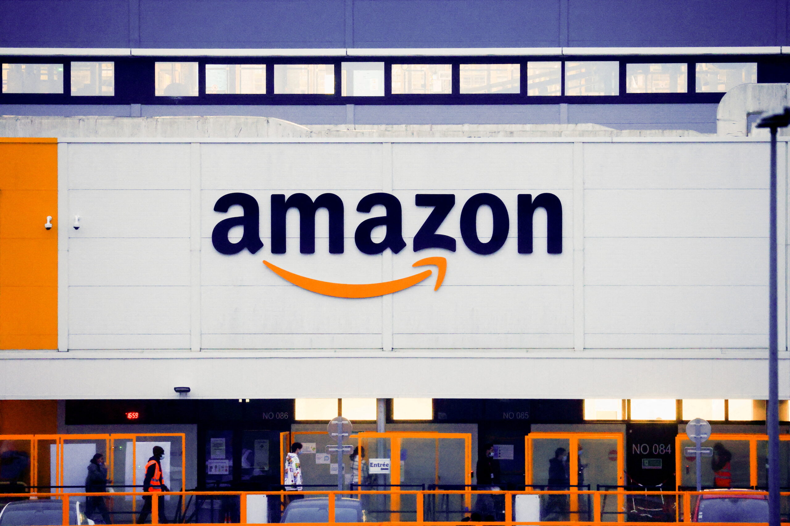 Exterior view of an Amazon fulfillment center with the Amazon logo displayed prominently on the building. The structure features a combination of yellow and white walls, glass windows, and sliding doors. A few people are seen walking near the entrance, perhaps wondering, "Is Amazon a good stock to buy right now? | 5StarsStocks