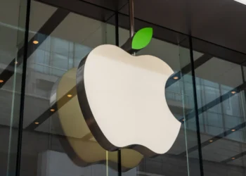 A large, white Apple logo with a green leaf on top is mounted on the exterior glass wall of a modern building, symbolizing the strength of Apple stock. The logo is prominently displayed, and the background shows reflections of the building's interior. | 5StarsStocks