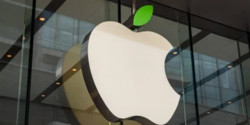 A large, white Apple logo with a green leaf on top is mounted on the exterior glass wall of a modern building, symbolizing the strength of Apple stock. The logo is prominently displayed, and the background shows reflections of the building's interior. | 5StarsStocks