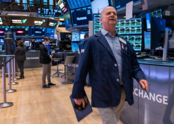 A person in a blue jacket walks through a bustling trading floor, holding a tablet. Multiple monitors displaying Dow Jones stocks and financial data are visible in the background, along with other people working. The environment is busy and well-lit. | 5StarsStocks