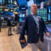A person in a blue jacket walks through a bustling trading floor, holding a tablet. Multiple monitors displaying Dow Jones stocks and financial data are visible in the background, along with other people working. The environment is busy and well-lit. | 5StarsStocks