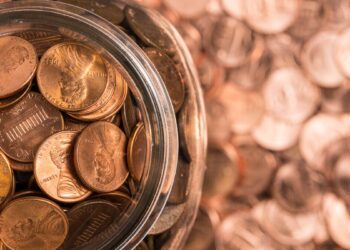A glass jar filled with US pennies sits on a surface also covered by numerous scattered pennies. The jar is open, revealing a mix of shiny and worn pennies, reminiscent of the fluctuations in penny stocks. The background is blurred, emphasizing the large quantity of coins. | 5StarsStocks