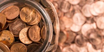 A glass jar filled with US pennies sits on a surface also covered by numerous scattered pennies. The jar is open, revealing a mix of shiny and worn pennies, reminiscent of the fluctuations in penny stocks. The background is blurred, emphasizing the large quantity of coins. | 5StarsStocks
