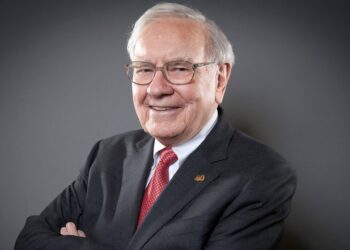 An elderly man with white hair and glasses, reminiscent of Warren Buffett, is smiling and wearing a dark suit with a white shirt and a red tie. He stands with his arms crossed against a plain, dark grey background. | 5StarsStocks