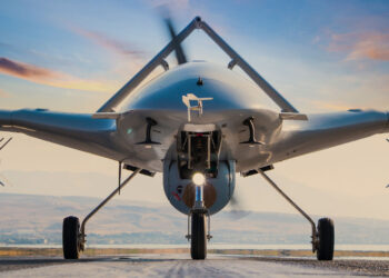 A front view of a military drone with a sleek, aerodynamic design, positioned on a runway. The sky in the background is painted with hues of orange and pink from the setting sun, creating a dramatic and picturesque scene—a testament to innovations impacting Aerospace & Defense Stocks. | 5StarsStocks