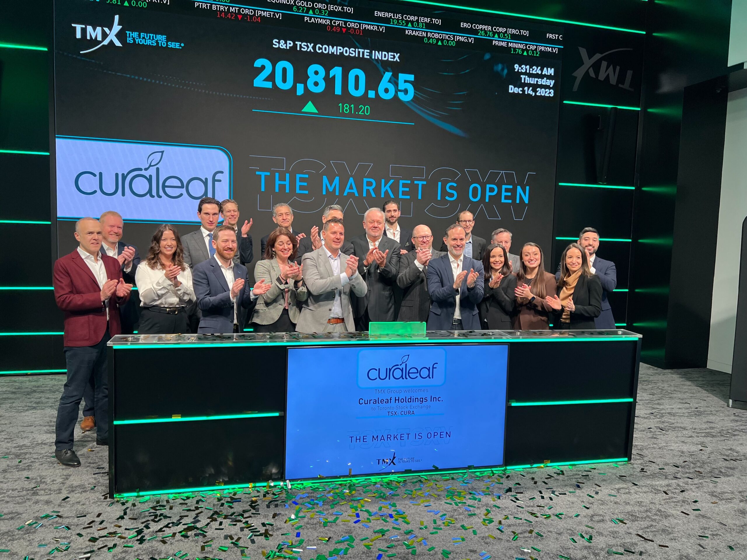 A group of people stands behind a podium at the Toronto Stock Exchange, clapping and smiling. The display board above shows "S&P TSX Composite Index 20,810.65" and a sign on the podium reads, "Curaleaf Holdings Inc. Curaleaf stock is up; the market is open. | 5StarsStocks