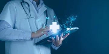 A medical professional in a white coat and stethoscope touches a glowing tablet with a holographic display of a healthcare cross and AI symbols, representing digital health technology, artificial intelligence, and the future potential of healthcare stocks. The background is dark and blurred. | 5StarsStocks