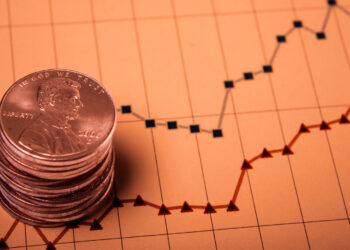A stack of U.S. pennies, symbolizing penny stocks, is placed on a financial graph with line charts. The graph features red and blue lines depicting financial trends over time, with grid lines and data points visible. The overall tone of the image is warm and slightly sepia. | 5StarsStocks