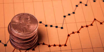 A stack of U.S. pennies, symbolizing penny stocks, is placed on a financial graph with line charts. The graph features red and blue lines depicting financial trends over time, with grid lines and data points visible. The overall tone of the image is warm and slightly sepia. | 5StarsStocks