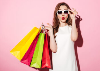 A woman in a white dress and white sunglasses holds colorful shopping bags in one hand while adjusting her sunglasses with the other. She has a surprised expression, reflecting the allure of consumer discretionary stocks, as she stands against a pink background. | 5StarsStocks
