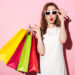 A woman in a white dress and white sunglasses holds colorful shopping bags in one hand while adjusting her sunglasses with the other. She has a surprised expression, reflecting the allure of consumer discretionary stocks, as she stands against a pink background. | 5StarsStocks