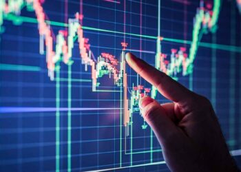 A hand points to a financial chart on a computer screen, indicating fluctuating stock market data with red and green candlestick patterns. The chart features multiple intersecting lines with price points, illustrating stocks trading trends and analysis. | 5StarsStocks