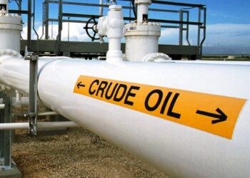 A large white pipeline labeled "CRUDE OIL" with black arrows pointing horizontally on a yellow sign. The pipeline is part of an industrial facility with various pipes, valves, and a metal structure in the background under a clear sky—an essential hub for driving oil stocks. | 5StarsStocks