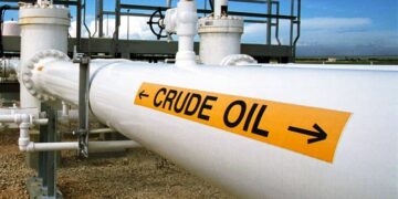 A large white pipeline labeled "CRUDE OIL" with black arrows pointing horizontally on a yellow sign. The pipeline is part of an industrial facility with various pipes, valves, and a metal structure in the background under a clear sky—an essential hub for driving oil stocks. | 5StarsStocks