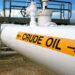 A large white pipeline labeled "CRUDE OIL" with black arrows pointing horizontally on a yellow sign. The pipeline is part of an industrial facility with various pipes, valves, and a metal structure in the background under a clear sky—an essential hub for driving oil stocks. | 5StarsStocks