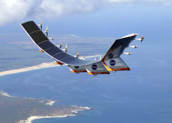 A solar-powered aircraft with multiple propellers and solar panels on its wings flies over a coastal landscape. The coastline features sandy beaches and lush greenery, while blue ocean waters extend into the horizon. The aircraft, resembling innovative designs like AVAV stock, has NASA logos on its body. | 5StarsStocks