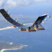 A solar-powered aircraft with multiple propellers and solar panels on its wings flies over a coastal landscape. The coastline features sandy beaches and lush greenery, while blue ocean waters extend into the horizon. The aircraft, resembling innovative designs like AVAV stock, has NASA logos on its body. | 5StarsStocks